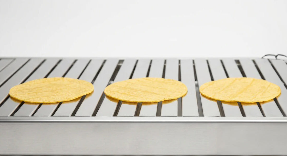 A polished stainless steel commercial tortilla oven slatbelt at side elevation, three fresh corn tortillas resting on the slats in a tidy row on a clean white studio background