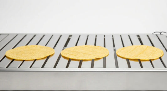 A polished stainless steel commercial tortilla oven slatbelt at side elevation, three fresh corn tortillas resting on the slats in a tidy row on a clean white studio background