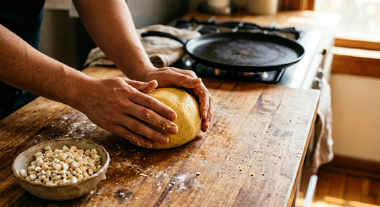 Two hands kneading a smooth golden ball of fresh masa dough on a wooden countertop, with a small bowl of nixtamalized corn kernels in the foreground and a cast iron comal griddle in soft focus behind